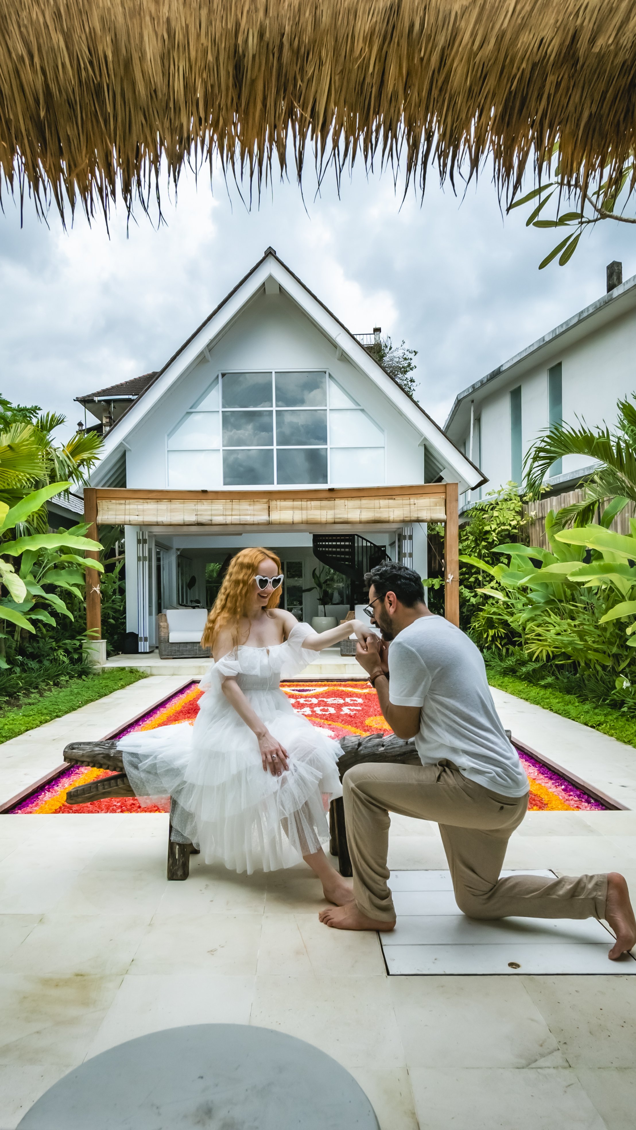 A man proposes to a woman sitting on a wooden bench outdoors in front of a modern white house with large windows, surrounded by lush green plants and a vibrant flower bed, on a cloudy day.