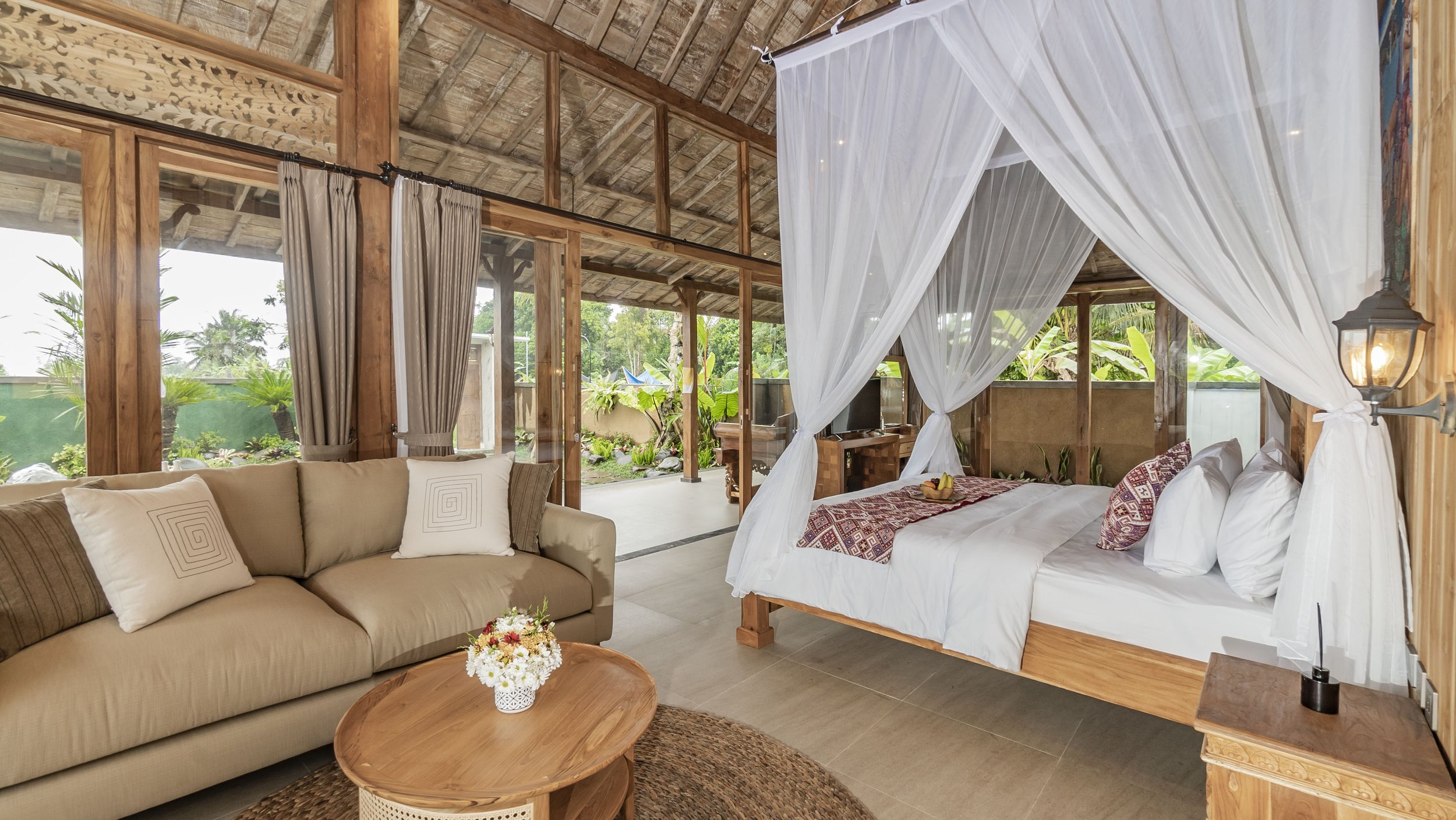 A tropical house with a tiled roof and large glass doors opens to a wooden patio with two lounge chairs by a small pool, surrounded by lush greenery and trees.