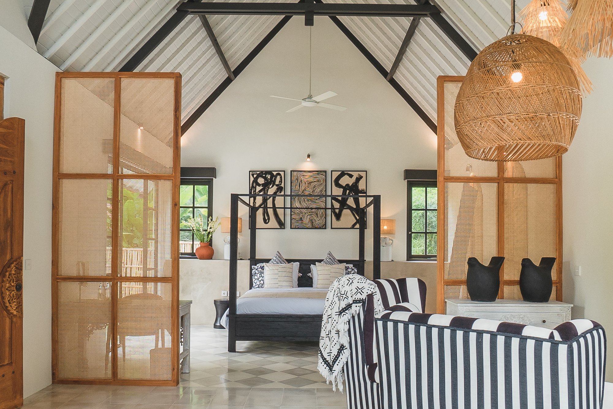 A spacious bedroom with high vaulted ceilings featuring exposed black beams, a large four-poster bed with black frame and decorative throw pillows, abstract black and white artwork above it, two windows with black framing on either side, and a stripe