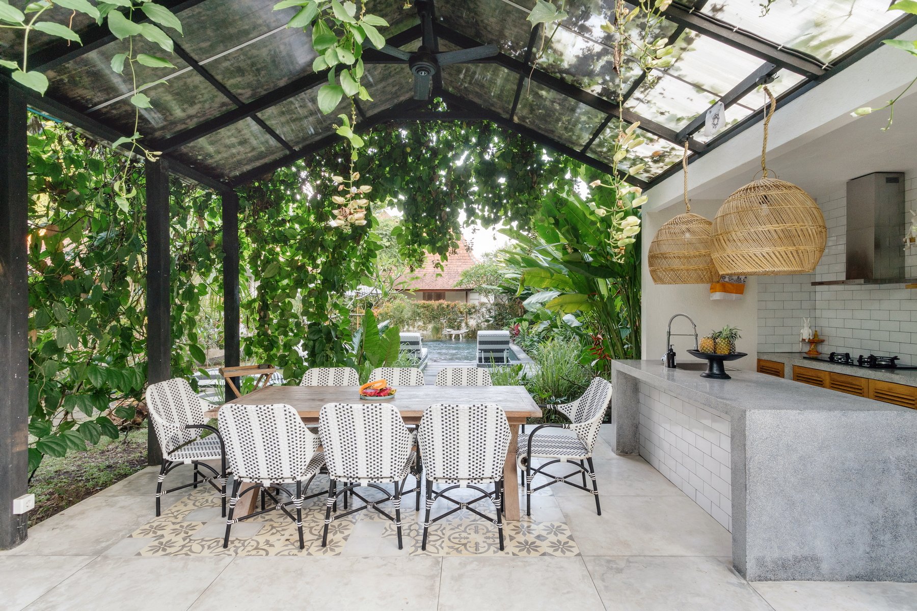 Outdoor dining area with a wooden table and ten white and black patterned chairs, lush green plants, and a kitchen counter with hanging wicker lamps in a semi-open space with a glass ceiling.