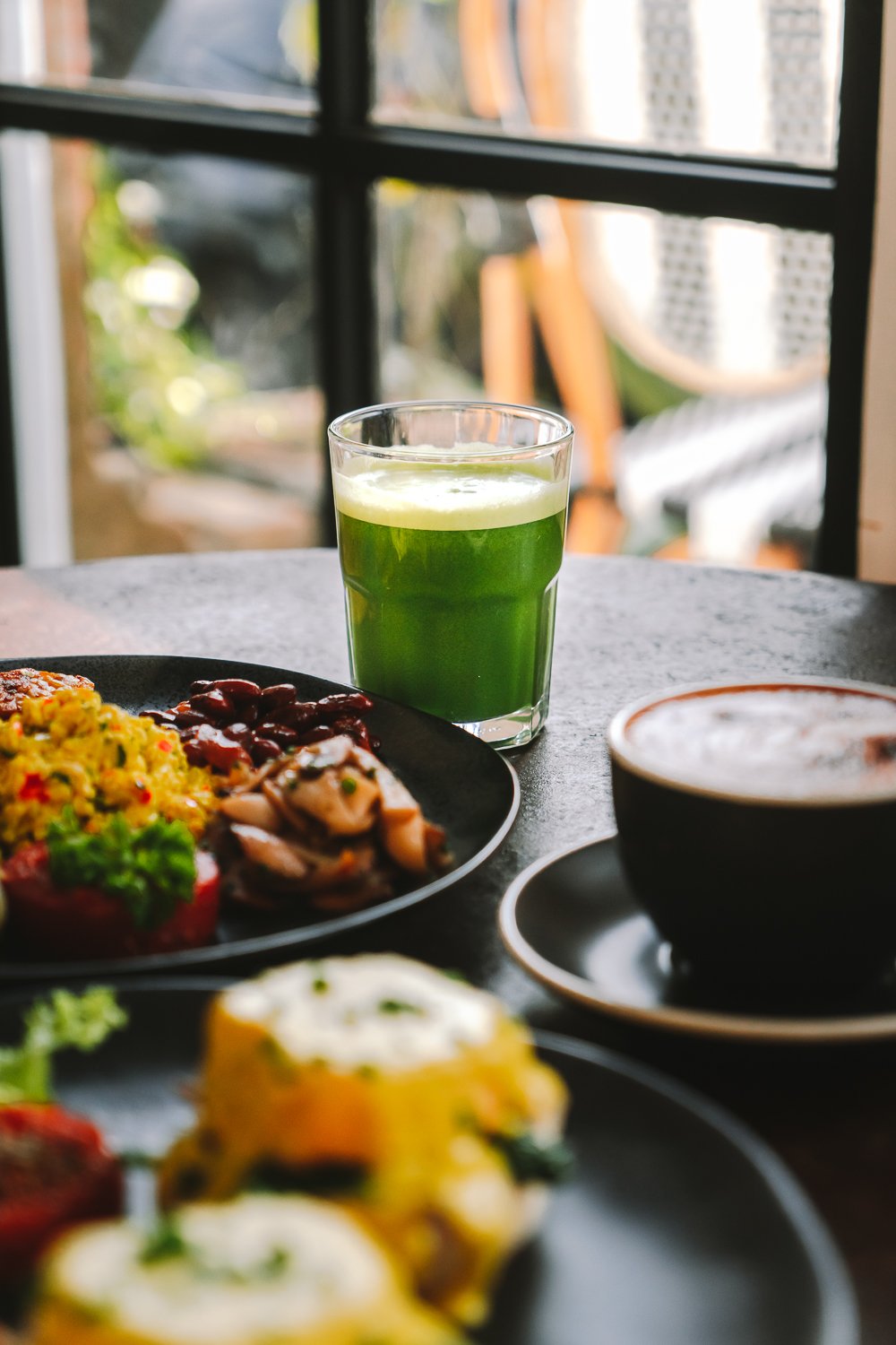 A glass of green juice, a cup of coffee, and a plate of assorted breakfast foods on a table near a window with outdoor scenery.