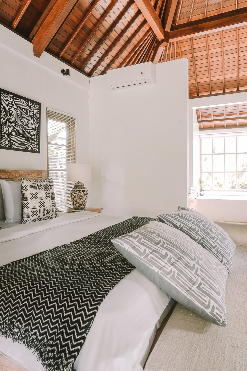 A bedroom with a white bed, patterned pillows, a black and white runner, a wooden headboard, a decorative lamp, and a window with blinds and natural light, featuring a wooden vaulted ceiling.