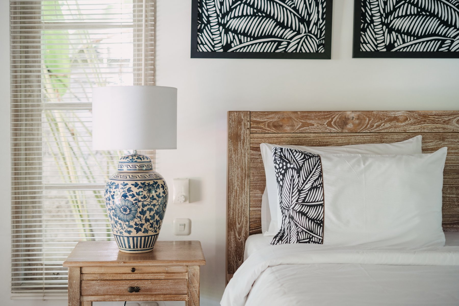 A bedroom featuring a wooden headboard, white bedding with black and white patterned pillows, a wooden nightstand with a blue and white floral ceramic lamp, a window with blinds, and black and white wall art.