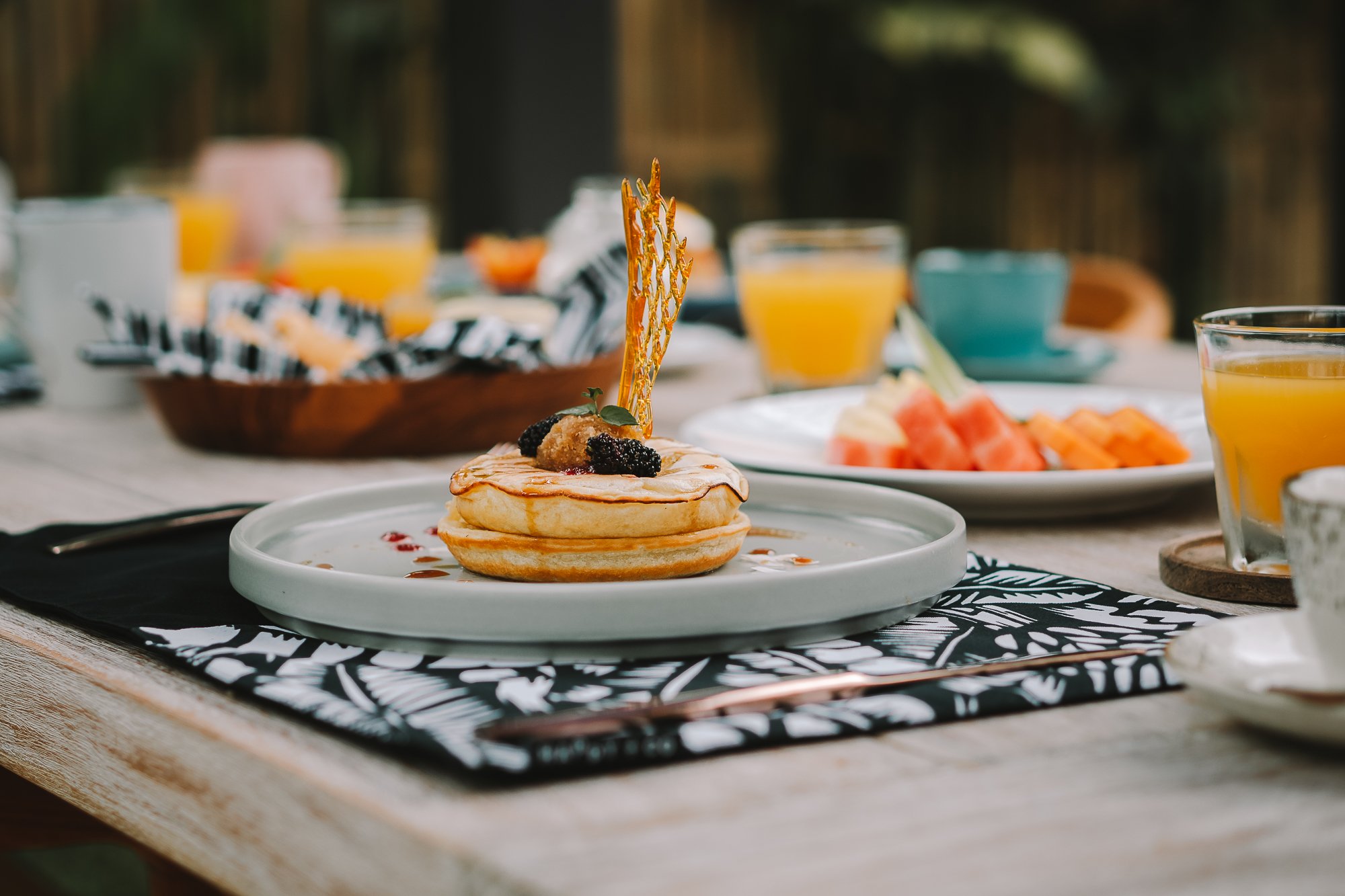A stack of pancakes topped with fruit and a caramel lattice on a white plate, with orange juice and fruit on the table, set outdoors.