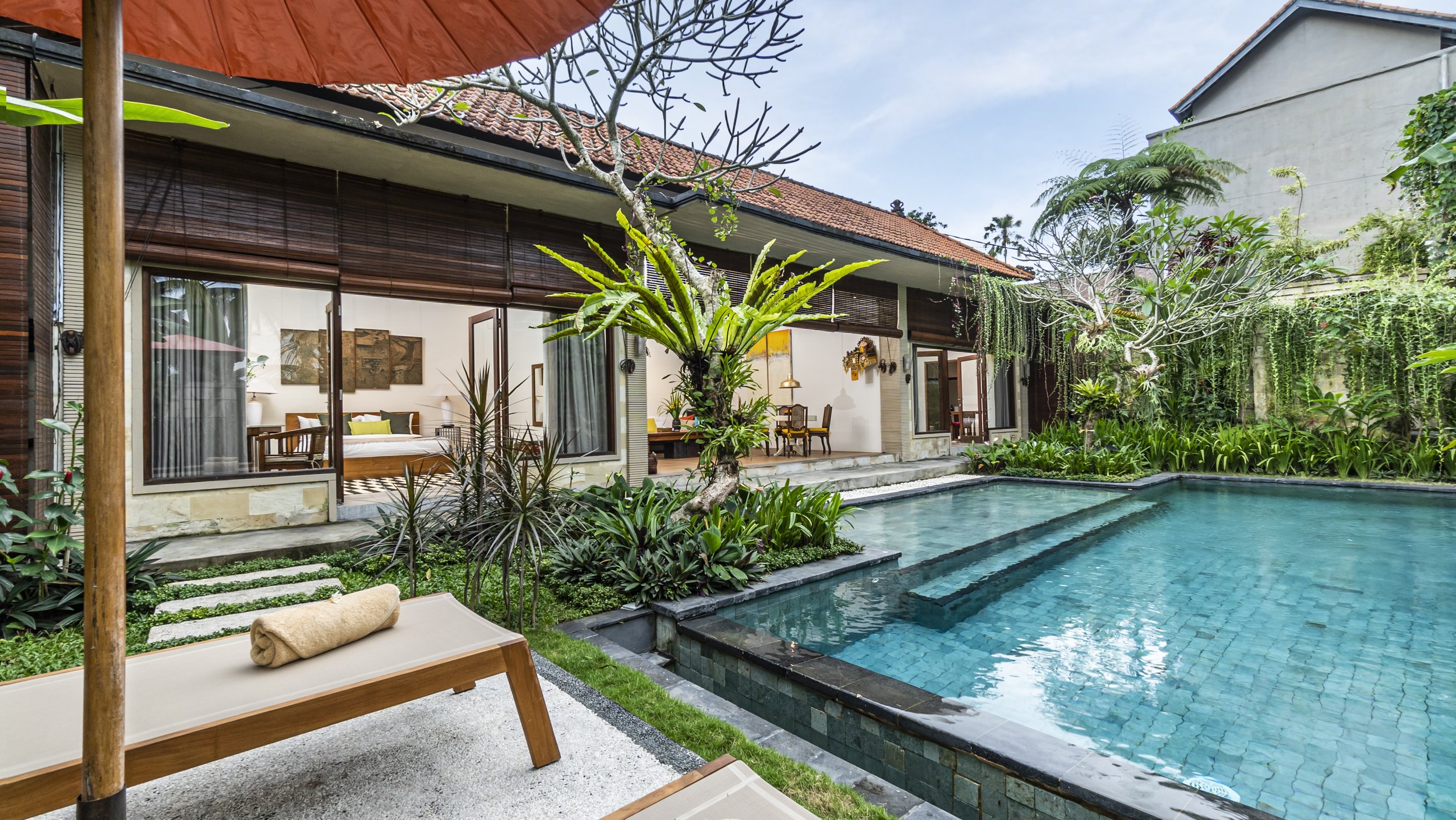 A tropical house with a tiled roof and large glass doors opens to a wooden patio with two lounge chairs by a small pool, surrounded by lush greenery and trees.